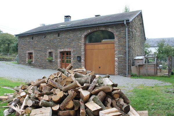 La ferme en pierre avec sa grande porte verte et une pile de bois de chauffage devant.