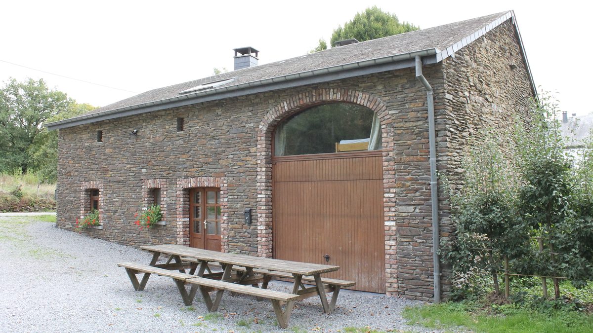 La ferme en pierre vue de côté, avec sa grande porte en arc et deux tables de pique-nique sur le gravier devant.